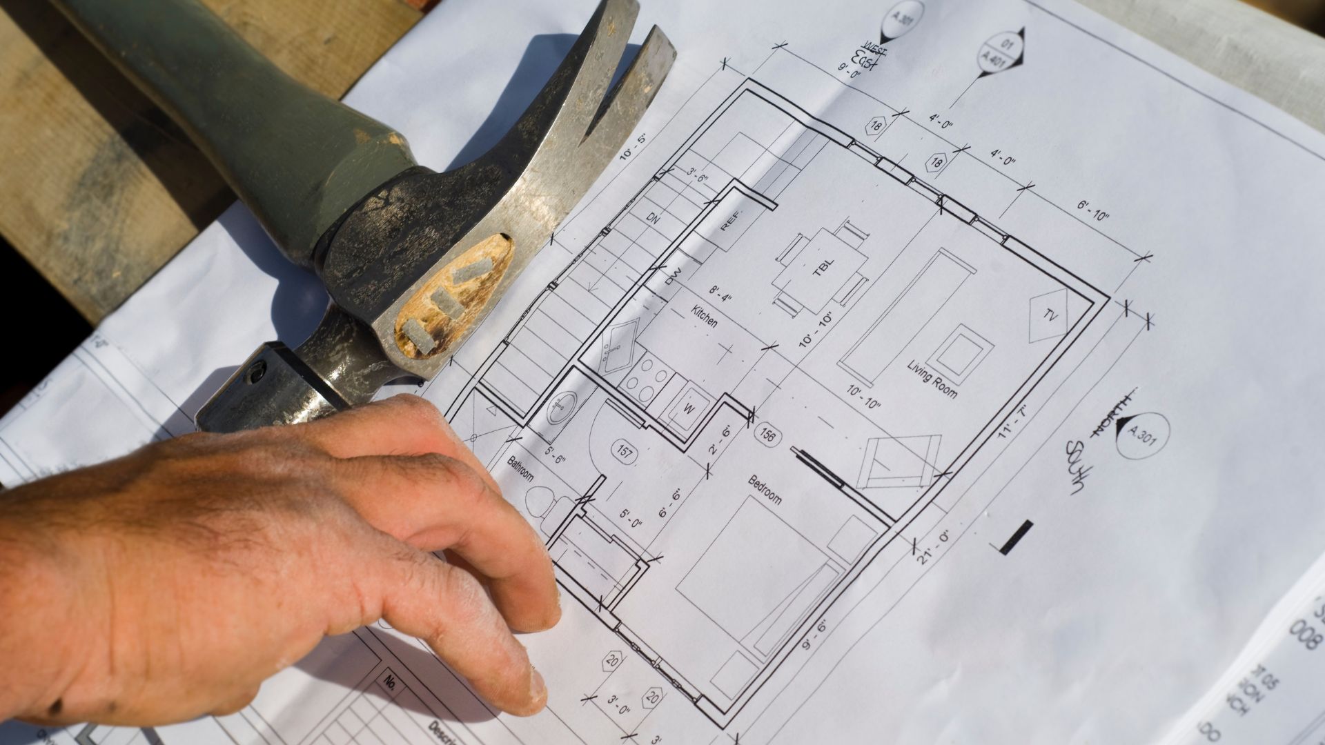 Person's hand next to a hammer pointing to a blueprint.